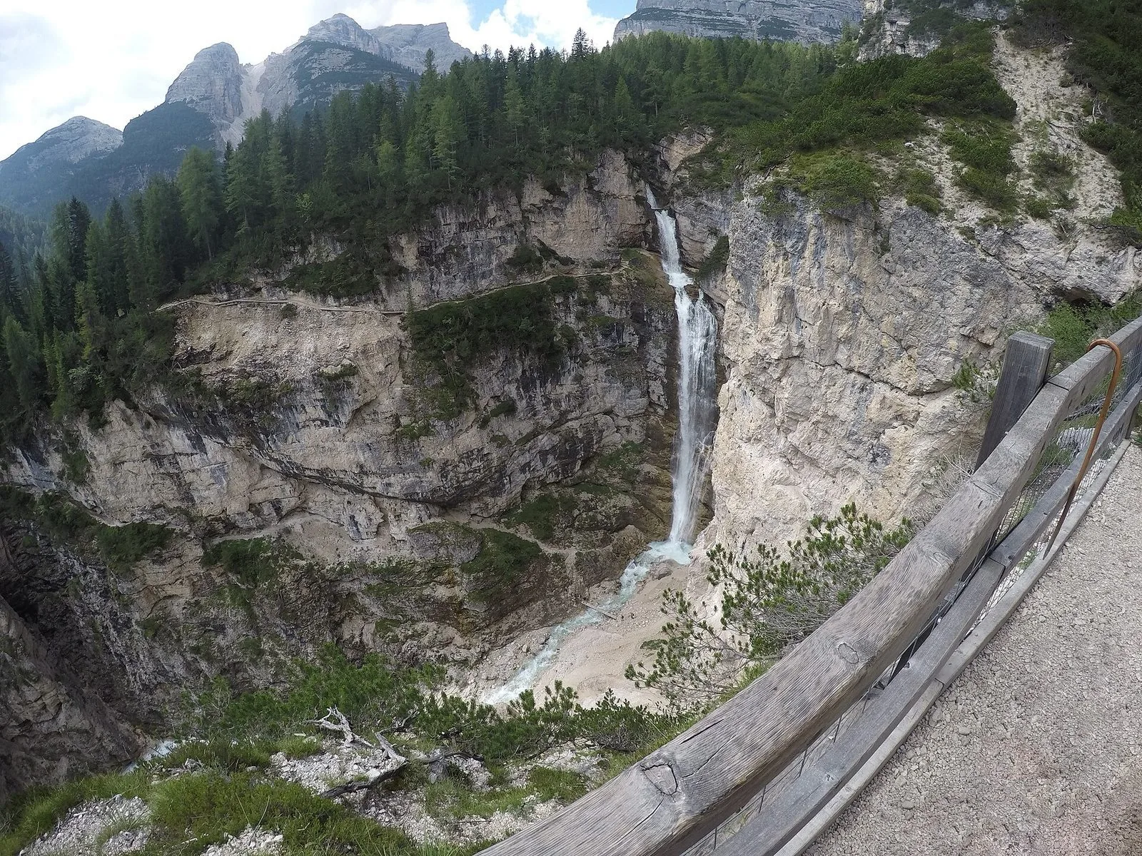 La cascata di Fanes che precipita in una forra rocciosa circondata da boschi di conifere, vista da un belvedere con staccionata in legno